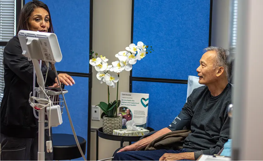 A healthcare worker speaks to a seated male patient in a medical clinic room with equipment and a white orchid on a side table.